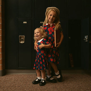 Posh Peanut Lilian Collared Twirl Dress Two children in matching red and blue dresses standing in front of a dark door.