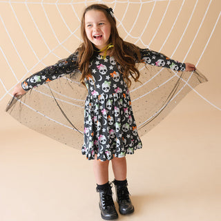 Posh Peanut Kelly Skelly Glow in the Dark Twirl Dress
Young girl wearing a colorful skull patterned dress with a beige background