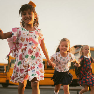 Posh Peanut Sabrina Criss-Cross Twirl Dress Three young girls walking together with a school bus in the background