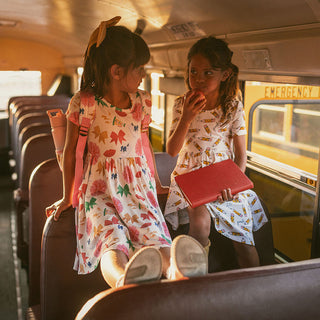Posh Peanut Sabrina Criss-Cross Twirl Dress Two young girls on a school bus, one holding a red book.