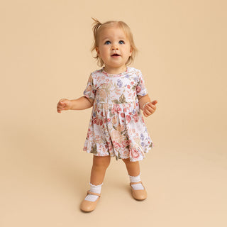 Posh Peanut Ruffled Twirl Bodysuit Dress - Kristiana
A toddler girl wearing a short-sleeve white dress with a pink, red, and green floral print, standing against a solid beige studio background.