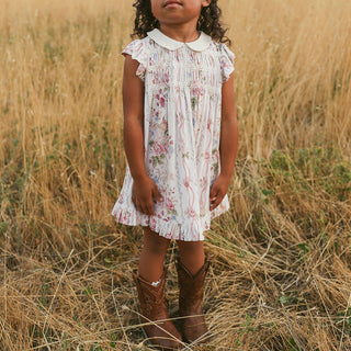 Posh Peanut Collared Smocked Ruffled Dress - Janey
A young girl wearing a white smocked dress with a pink floral print and a white Peter Pan collar, standing in a field of dry golden grass and wearing brown cowboy boots.