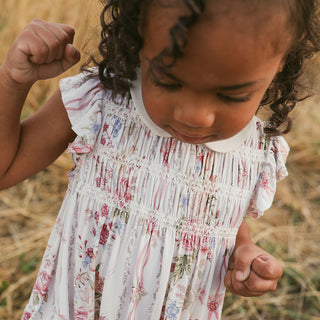 Posh Peanut Collared Smocked Ruffled Dress - Janey
A toddler girl wearing a white smocked dress with a pink and blue floral print, photographed outdoors in a dry grassy field.