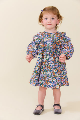 Pink Chicken Baby Girls Everly Dress Set - True Blue Sketched Floral
A toddler girl with light brown hair wearing a blue floral long-sleeve dress and dark gray shoes, standing against a solid beige background.