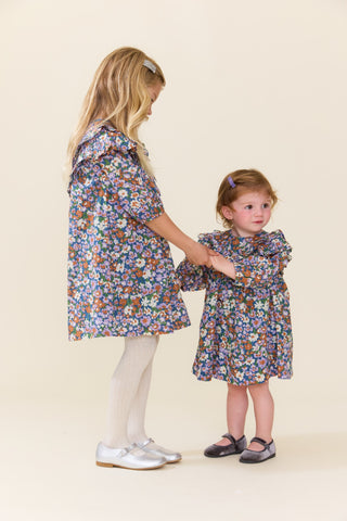 Pink Chicken Baby Girls Everly Dress Set - True Blue Sketched Floral
Two young girls modeling matching floral print dresses in a studio setting. The older girl has blonde hair and wears white tights and silver shoes, while the younger girl has light brown hair and wears dark shoes.