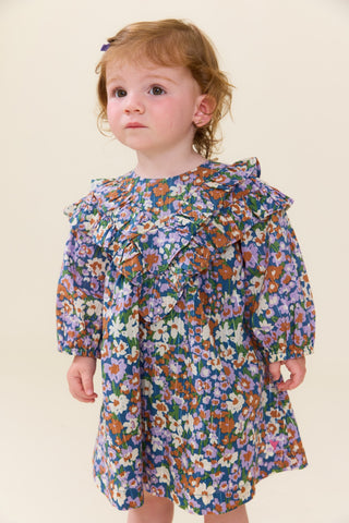 Pink Chicken Baby Girls Everly Dress Set - True Blue Sketched Floral
A toddler girl with light brown hair wearing a blue floral long-sleeve dress with ruffles, standing against a plain beige background.