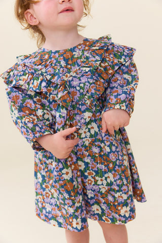 Pink Chicken Baby Girls Everly Dress Set - True Blue Sketched Floral 
A toddler wearing a long-sleeve floral romper with a ruffled neckline, photographed against a plain cream background.