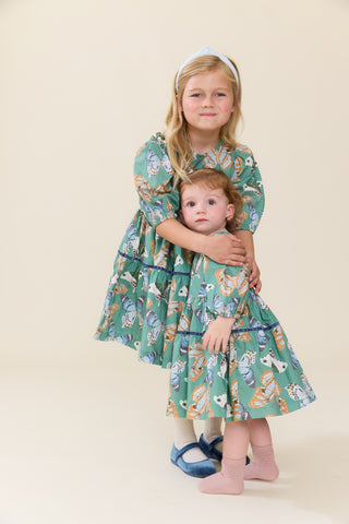 Pink Chicken Baby Girls Maribelle Dress Set - Green Butterflies
Two young girls wearing matching green tiered dresses with a whimsical animal print, standing against a solid beige background. The older girl has blonde hair and wears a white headband, while the younger girl has light brown hair and wears pink socks.