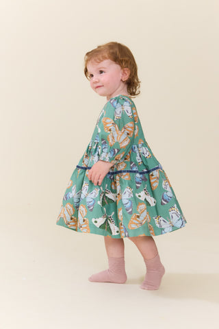 Pink Chicken Baby Girls Maribelle Dress Set - Green Butterflies
A toddler girl with curly light brown hair is wearing a tiered green dress with a colorful butterfly and owl pattern, paired with pink socks, standing against a solid cream background.