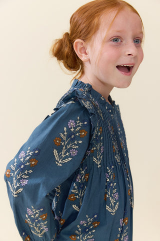Pink Chicken Girls Jaipur Dress - Balsam Dewdrop
A young girl with red hair tied back in a low bun is shown in profile wearing a blue long-sleeve blouse with a floral print. The blouse features a smocked bodice and ruffled details.