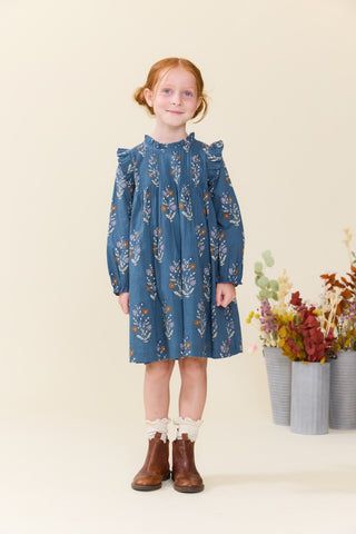Pink Chicken Girls Jaipur Dress - Balsam Dewdrop
A young girl with red hair in a ponytail stands wearing a blue long-sleeve dress with a white and orange floral pattern, paired with white ruffled socks and brown boots. She is posed in front of a warm cream background with decorative dried flowers in gray pots on the right.