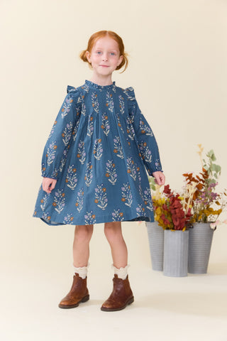 Pink Chicken Girls Jaipur Dress - Balsam Dewdrop
A young girl with red hair stands wearing a blue long-sleeve dress with a white and orange floral pattern, paired with white socks and brown boots, in front of a beige background with decorative flower pots.