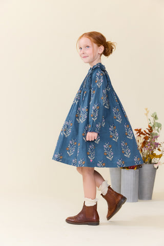 Pink Chicken Girls Jaipur Dress - Balsam Dewdrop
A young girl with red hair tied back wears a blue long-sleeve dress with a white and orange floral pattern, paired with brown ankle boots and white socks, standing in a studio setting with decorative potted plants in the background.