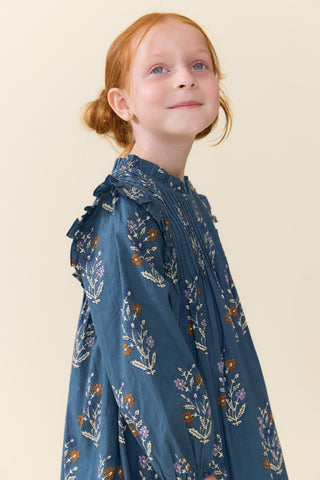 Pink Chicken Girls Jaipur Dress - Balsam Dewdrop 
A young girl with red hair tied back wears a blue long-sleeve dress with a delicate white and orange floral pattern, photographed against a solid light beige background.