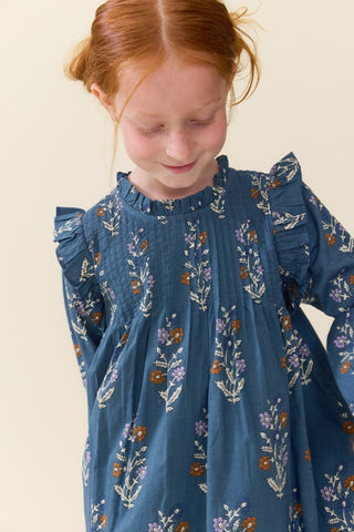 Pink Chicken Girls Jaipur Dress - Balsam Dewdrop
A young girl with red hair looking down, wearing a blue long-sleeve dress with a white and purple floral pattern, set against a plain beige background.