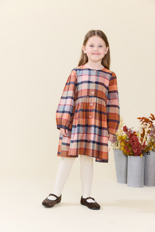 Pink Chicken Girls Mae Dress - Hayride Plaid
A young girl stands against a warm cream studio backdrop wearing a long-sleeve plaid dress in orange, pink, navy, and beige tones, paired with white tights and dark patterned shoes. Decorative flower pots sit to the right.