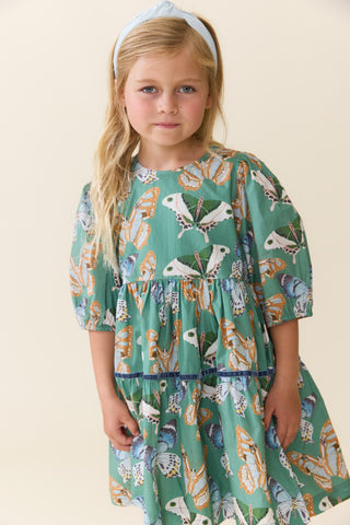 Pink Chicken Girls Maribelle Dress - Green Butterflies
A young girl with blonde hair wearing a light blue headband and a green tiered dress with a butterfly and bird pattern, standing against a plain beige background.