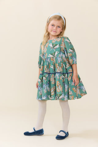 Pink Chicken Girls Maribelle Dress - Green Butterflies
A young girl with blonde hair in a ponytail stands against a solid cream background, wearing a green tiered dress with a colorful bird pattern, white tights, and navy blue shoes.