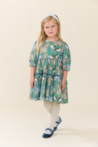 Pink Chicken Girls Maribelle Dress - Green Butterflies
A young girl with blonde hair wearing a tiered green dress with a colorful bird pattern, cream tights, and blue Mary Jane shoes, standing against a solid beige studio background.