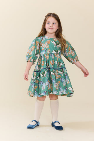 Pink Chicken Girls Maribelle Dress - Green Butterflies
A young girl with long light brown hair stands wearing a green tiered dress with a colorful butterfly and floral print, paired with cream knee-high socks and blue shoes, against a solid beige background.