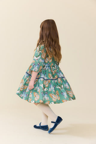 Pink Chicken Girls Maribelle Dress - Green Butterflies
A young girl with long light brown hair is shown from behind wearing a green tiered dress with a colorful butterfly print, white tights, and blue shoes, set against a solid beige background.