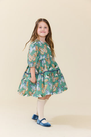 Pink Chicken Girls Maribelle Dress - Green Butterflies 
A young girl with long light brown hair stands in a studio wearing a tiered green dress with a colorful animal print, cream knee-high socks, and blue Mary Jane shoes against a solid beige background.