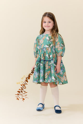 Pink Chicken Girls Maribelle Dress - Green Butterflies
A young girl stands against a warm cream studio background wearing a teal green dress with a whimsical multicolor print of animals and nature motifs. She is styled with white knee-high socks and blue Mary Jane shoes, holding a small decorative branch.