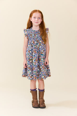 Pink Chicken Girls Stevie Dress - True Blue Sketched Floral
A young girl with long red hair stands against a plain beige studio background wearing a sleeveless, ruffled-sleeve floral dress with brown boots.