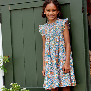 Pink Chicken Girls Stevie Dress - True Blue Sketched Floral
A young girl stands smiling in front of a dark green wooden door, wearing a colorful floral smocked dress with ruffled sleeves, white socks, and leopard-print boots.