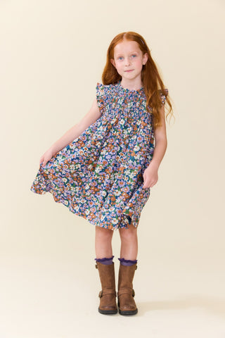Pink Chicken Girls Stevie Dress - True Blue Sketched Floral
A young girl with long red hair stands against a plain cream background wearing a sleeveless, ruffled-sleeve floral dress with brown boots.