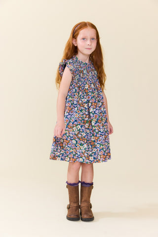 Pink Chicken Girls Stevie Dress - True Blue Sketched Floral 
A young girl with red hair stands against a solid beige background wearing a sleeveless, smocked floral dress with brown boots.