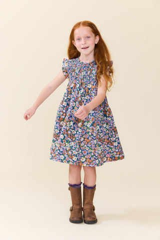 Pink Chicken Girls Stevie Dress - True Blue Sketched Floral
A young girl with long red hair stands against a plain beige studio background wearing a sleeveless knee-length dress with a dense multicolor floral print and brown boots.