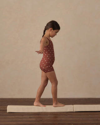 Play by Rylee & Cru Malibu Bodysuit - Brick Hearts
A young girl in a side profile stands on a gymnastics tumbling mat, wearing a rust-red sleeveless leotard with white polka dots against a textured beige wall and dark wood floor.