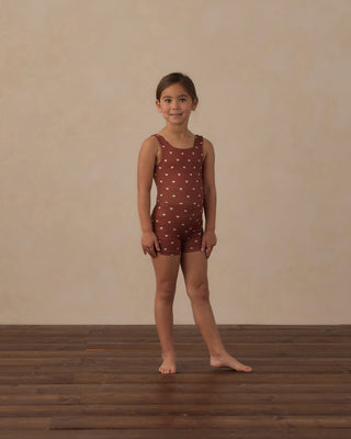 Play by Rylee & Cru Malibu Bodysuit - Brick Hearts
A young girl stands barefoot on a dark wood floor against a textured beige wall, wearing a sleeveless rust-brown one-piece swimsuit with a white polka-dot pattern.