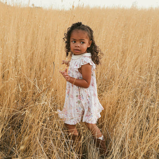 Posh Peanut Collared Smocked Ruffled Dress - Janey
A young girl with curly brown hair stands in a field of dry golden grass, wearing a white sleeveless dress with a pink and green floral pattern and a white mock neck.