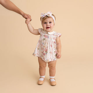 Posh Peanut Collared Smocked Ruffled Dress Bummie Set - Janey
A baby girl wearing a floral print romper, matching bow headband, and Mary Jane shoes stands while holding an adult's hand against a solid beige background.