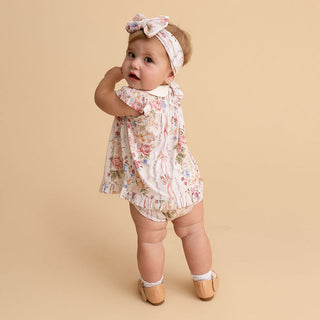 Posh Peanut Collared Smocked Ruffled Dress Bummie Set - Janey
A baby girl wearing a cream-colored floral romper with a ruffled hem, a matching floral bow headband, and tan leather shoes, standing against a solid beige studio background.