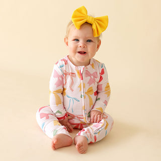 Posh Peanut Convertible One Piece - Shawna
Baby wearing a colorful outfit with a yellow bow on a beige background