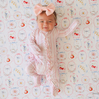 Posh Peanut Footie Ruffled Zippered One Piece - Remi
A baby lying on a patterned blanket, wearing a light pink footed sleeper with ruffle trim and a matching pink bow headband.