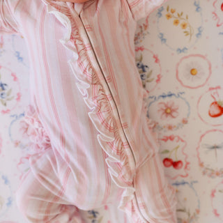 Posh Peanut Footie Ruffled Zippered One Piece - Remi
A close-up of a baby wearing a light pink and white striped footless bodysuit with a ruffled trim down the front, lying on a floral-patterned blanket.