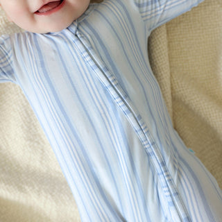 Posh Peanut Footie Zippered One Piece - Richie
A close-up of a baby wearing a light blue and white striped footless sleep sack, lying on a textured beige blanket.