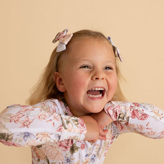 Posh Peanut Mini Bow Hair Clips - Kristiana 
A young girl with blonde hair styled with two matching floral hair bows, wearing a white long-sleeve top with a pink and green floral pattern, posing against a solid beige background.