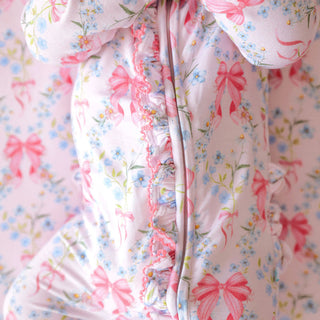 Posh Peanut Picot Footie Ruffled Zippered One Piece - Harriet
A close-up of a baby's foot wearing a light pink footed sleeper with a delicate blue floral print, pink bows, and ruffled trim along the front zipper.