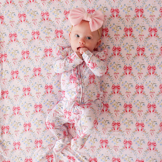 Posh Peanut Picot Footie Ruffled Zippered One Piece - Harriet
A baby lying on a pink floral blanket with bow patterns, wearing a matching pink footed sleeper and a large pink bow headband.