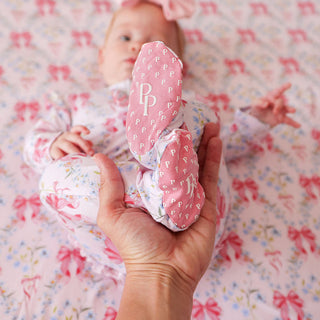 Posh Peanut Picot Footie Ruffled Zippered One Piece - Harriet
A baby wearing a pink and white floral romper lies on a matching pink floral blanket while an adult hand holds the baby's feet, which are wearing pink booties with a repeating monogram pattern and a large 'PP' monogram on the sole.