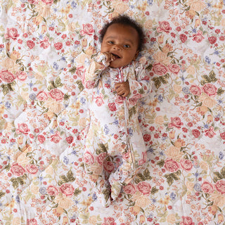 Posh Peanut Picot Footie Ruffled Zippered One Piece - Kristiana
A baby lying on a floral quilted blanket, wearing a matching footed swaddle or sleeper with a pink, peach, green, and blue floral print on a white background.