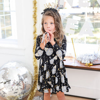 Posh Peanut Ruffle Twirl Dress - Midnight Bash
Young girl in a black dress with white patterns, standing in a festive room with decorations.