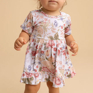 Posh Peanut Ruffled Twirl Bodysuit Dress - Kristiana
A toddler girl wearing a short-sleeve white dress with a pink, red, and green floral print, standing against a solid beige background.