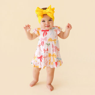 Posh Peanut Ruffled Twirl Bodysuit Dress - Shawna
Baby wearing a colorful dress with bow patterns and a yellow headband on a beige background
