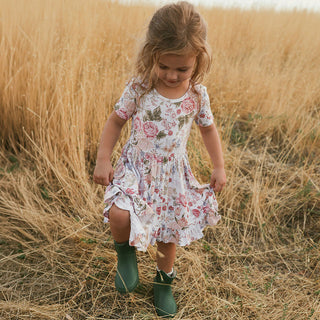 Posh Peanut Ruffled Twirl Dress - Kristiana
A young girl wearing a short-sleeve floral print dress and green rain boots stands in a field of dry golden grass, holding out the skirt of the dress.